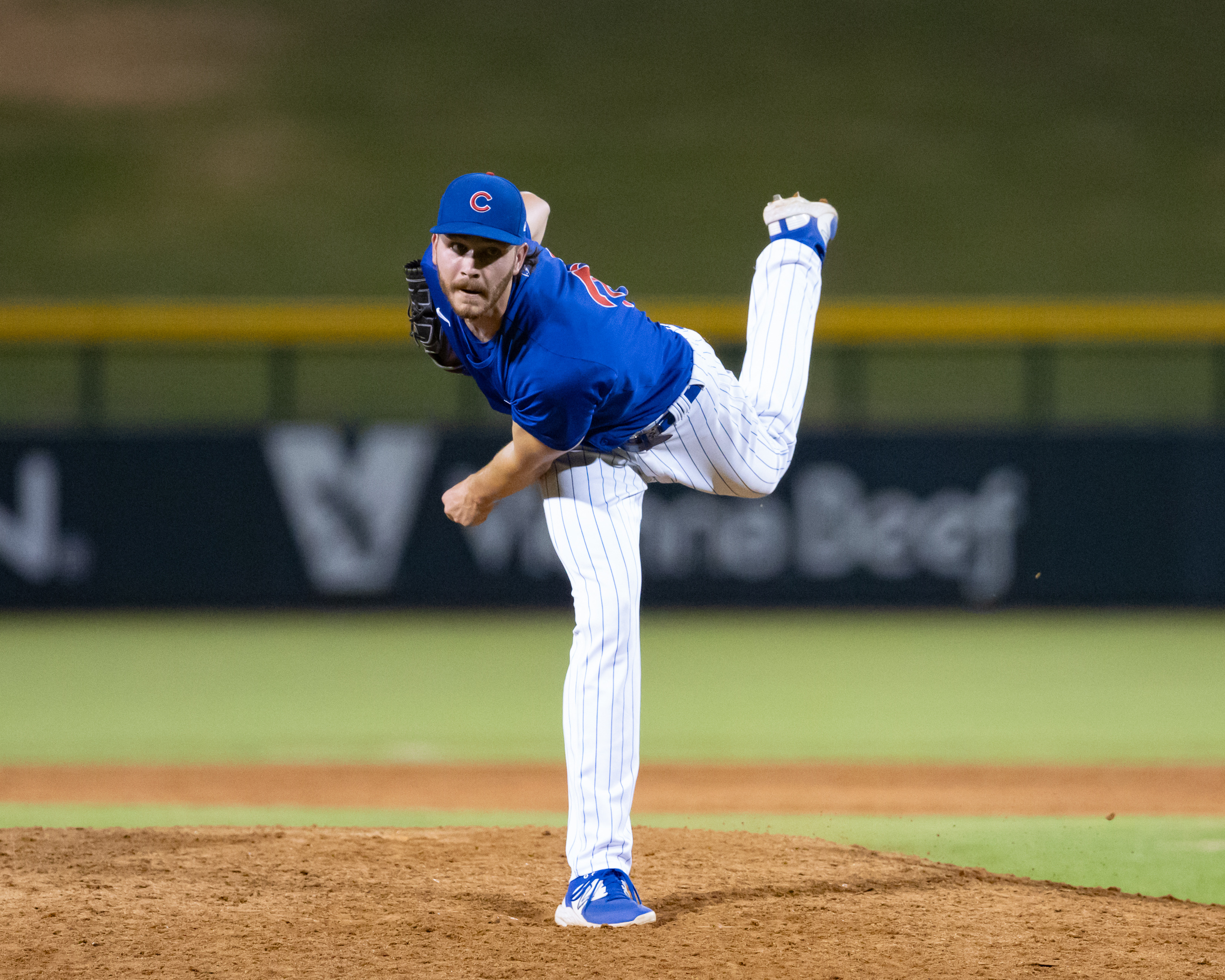 Wilson Cunningham pitching for the Cubs, mid-throw with leg lifted and focused expression, on a baseball field at night