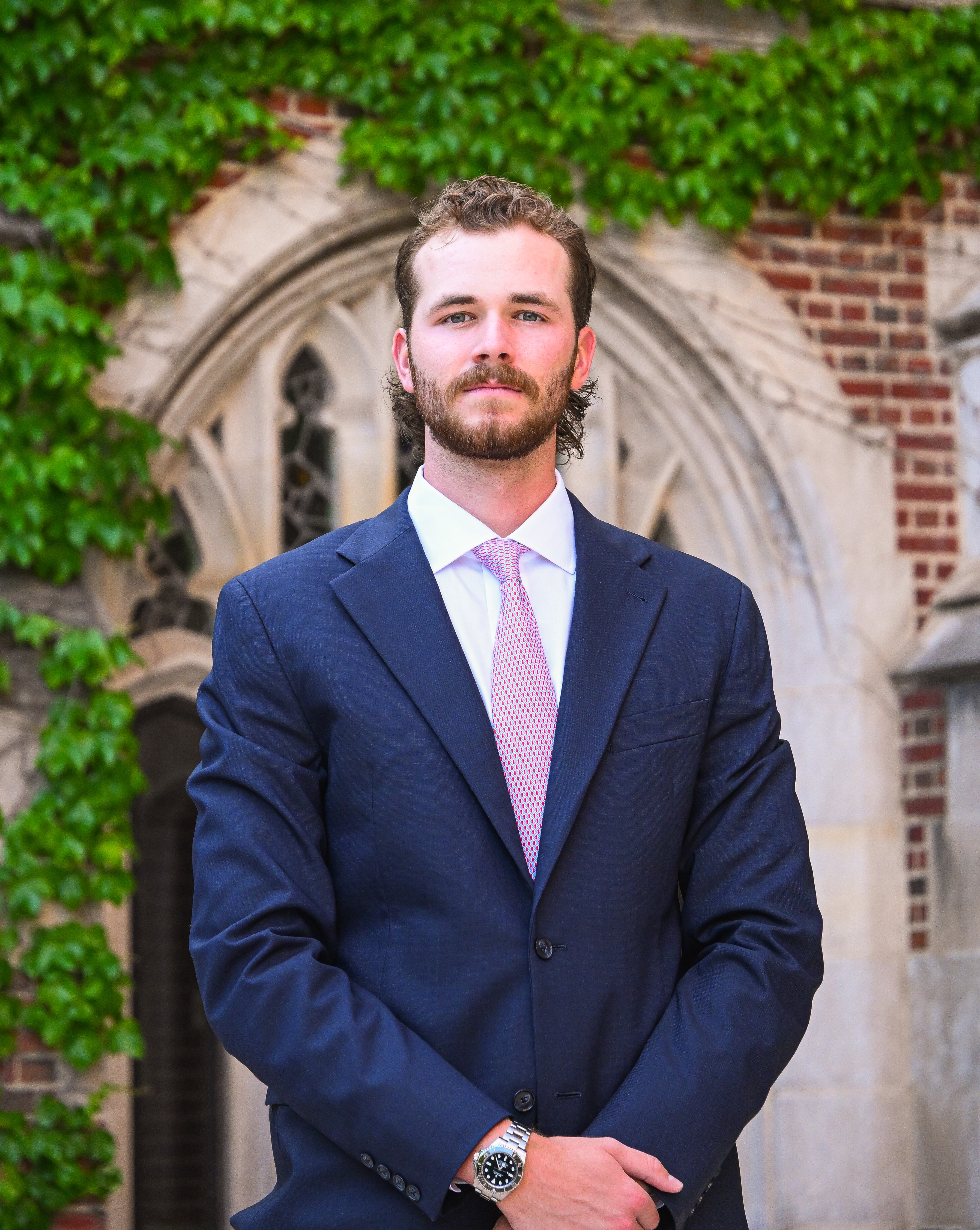 Wilson Cunningham standing in a navy blue suit and pink tie, posing confidently in front of a gothic archway with ivy-covered brick walls
