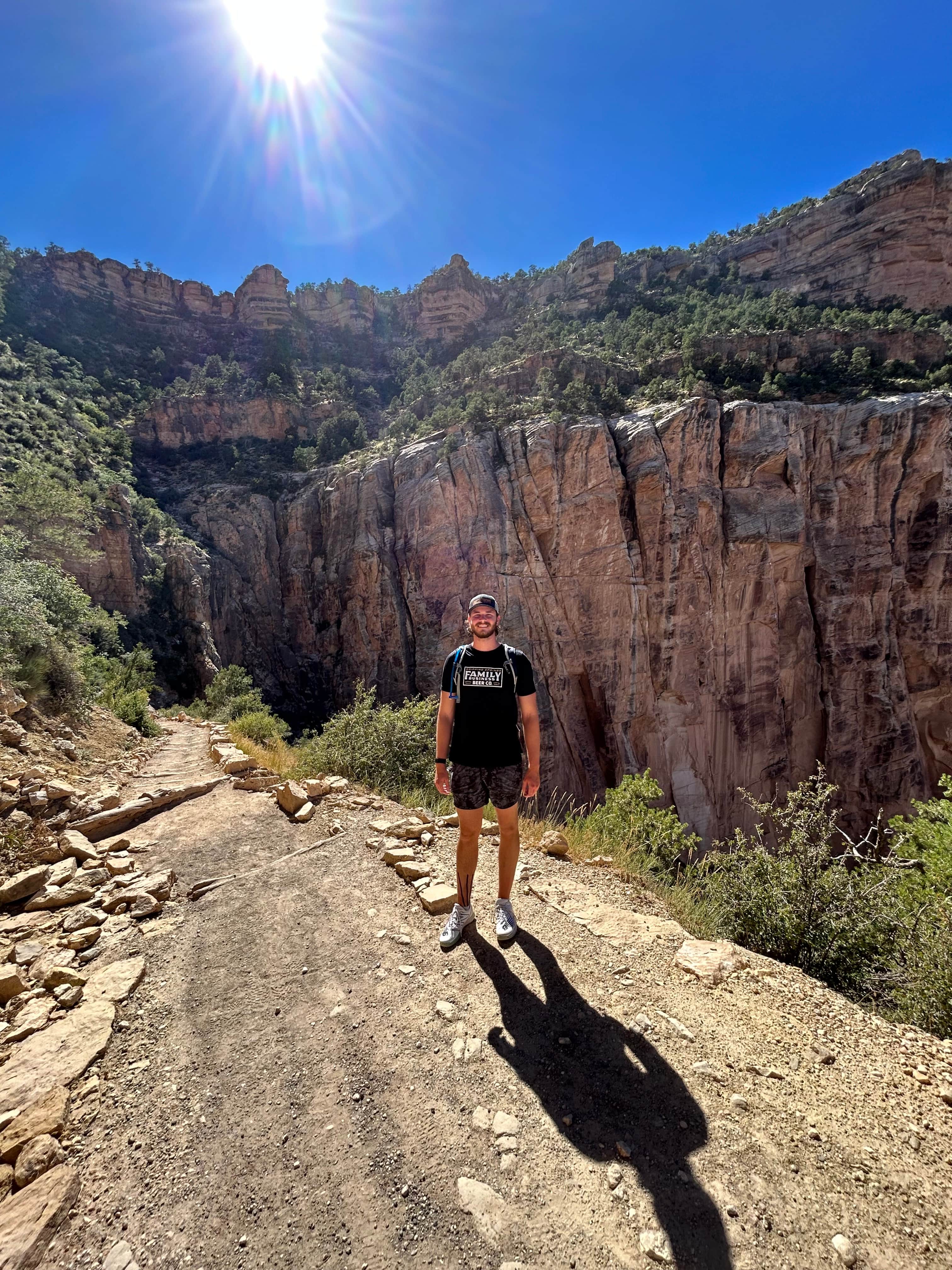 Hiker standing on sunlit trail with dramatic canyon cliffs and bright blue sky on Bright Angel Trail, Grand Canyon