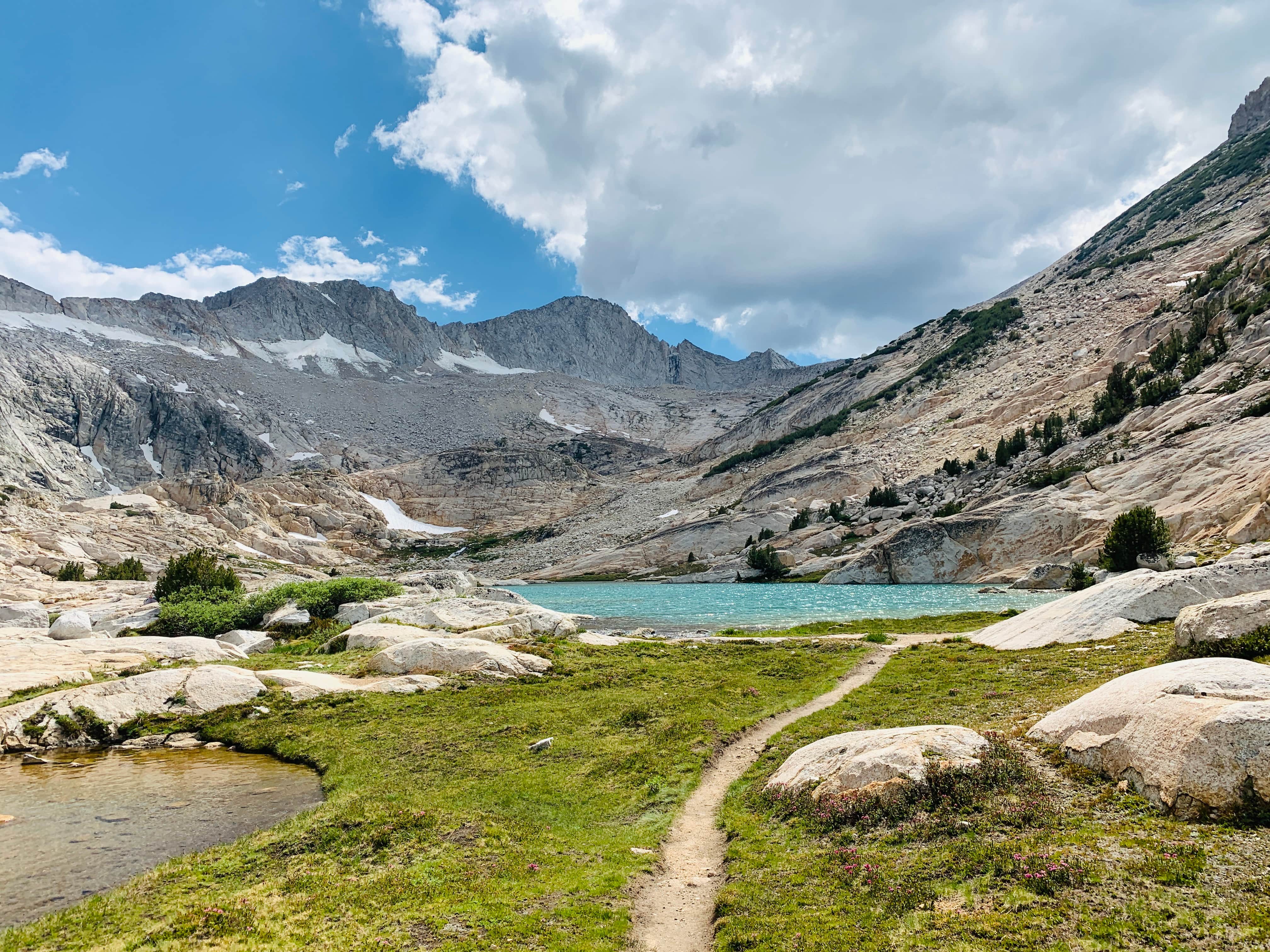 Mountain landscape with a rocky alpine trail leading to clear blue Conness Lake under a partly cloudy sky
