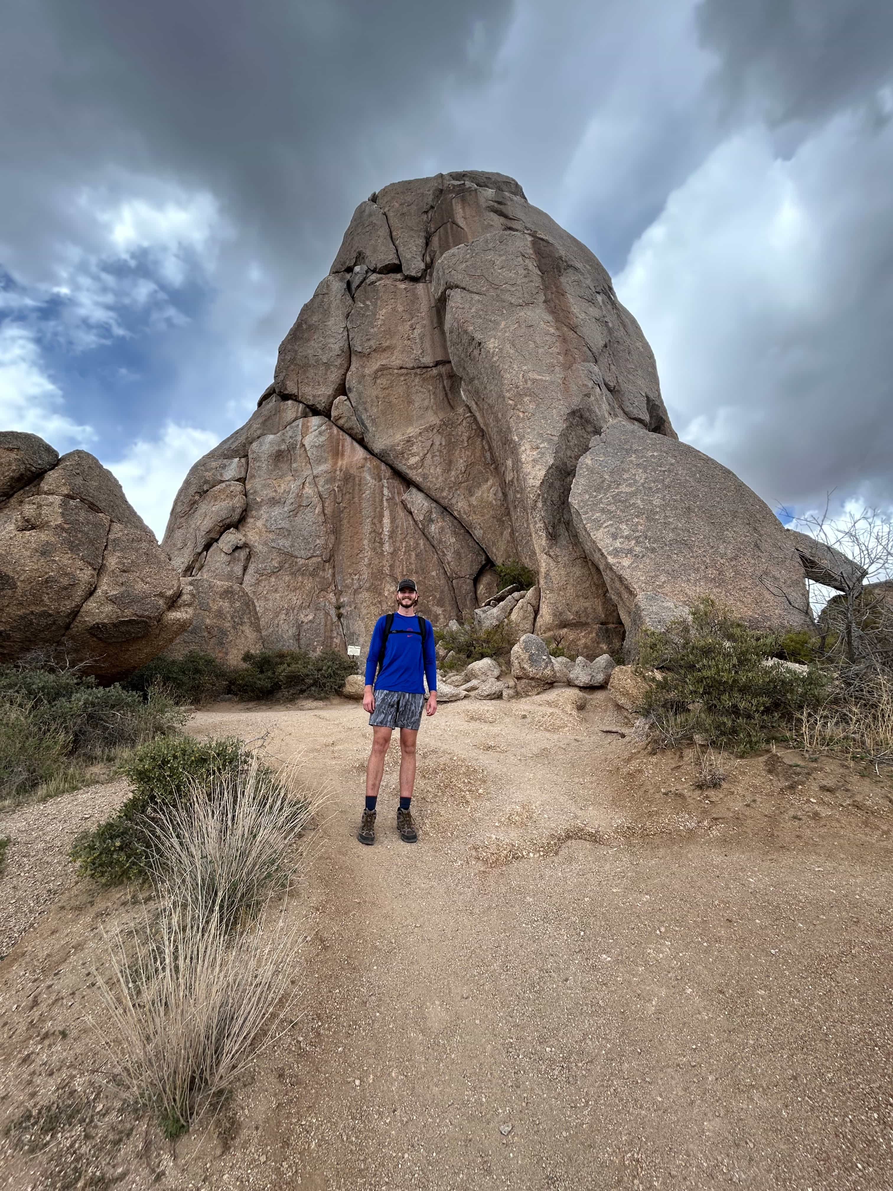 Hiker standing on sandy trail in front of large granite spire under dramatic clouds at Tom’s Thumb, Arizona